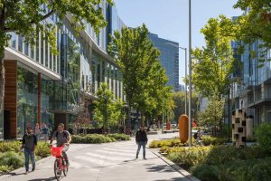 Urban street scene in the heart of Denny Triangle, with people walking and a cyclist on a pathway. Modern buildings and green trees line the street under Seattle's clear blue sky, offering a perfect backdrop for your tour.