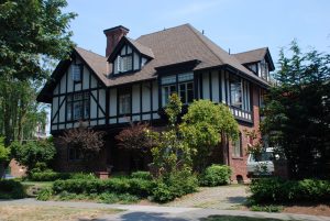 Explore a Tudor-style house with brown and white timber framing, brick accents, and multiple gables nestled among trees and shrubs on the Capitol Hill Tour.