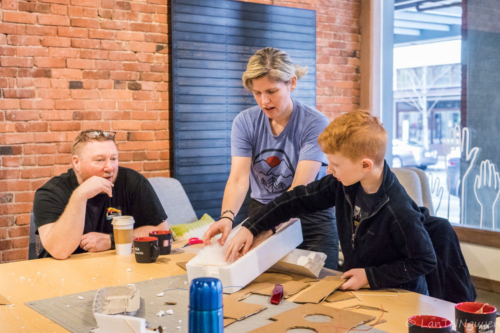 Two adults and a child immerse themselves in a family architectural workshop at the table, surrounded by cardboard and Styrofoam materials for their creative craft project.