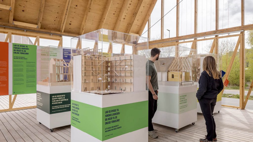 Two adults view architectural models on white platforms.