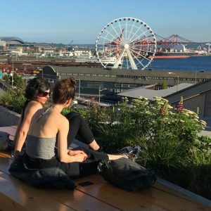 Two women sitting on the steps of overlook walk around sunset. The ferris wheel is visible in the distance. 