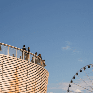 A close-up image of several people standing at the edge of overlook walk. Part of the Seattle ferris wheel is visible on the right. The sky is blue with some clouds.