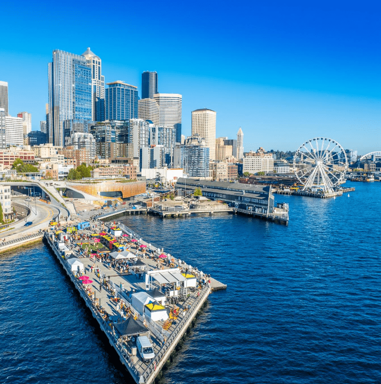 The Seattle waterfront on a sunny day. There are people on the pier and the Seattle ferris wheel and several skyscrapers are visible in the background.