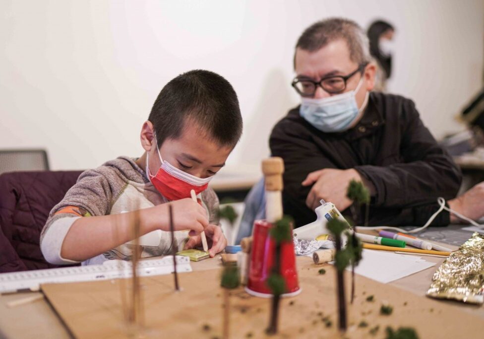 In a charming scene of creativity, a family engages in an architectural workshop. A child and an adult, both wearing masks, collaborate on crafting intricate models and trees laid out on the table.