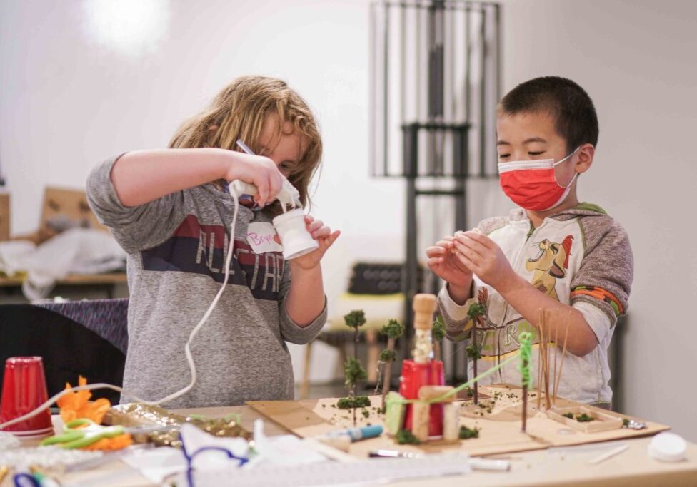 Two children engaged in a crafting project; one uses a hot glue gun, while the other, wearing a red mask, concentrates on an item. A cardboard artwork with small structures is on the table.