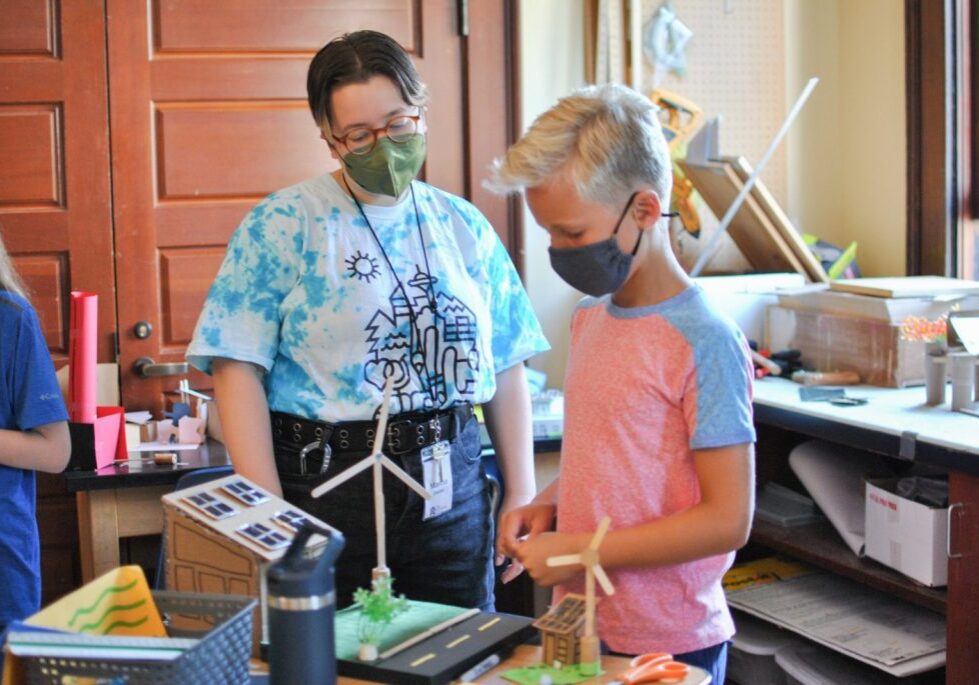 Two educators wearing masks work on an architectural model with wind turbines in a room filled with art supplies and projects.