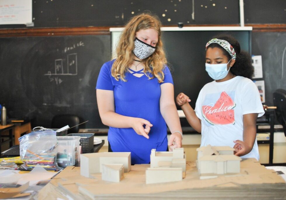 In a classroom alive with creativity, two students wearing masks focus intently on their scale model project. Surrounded by school supplies and a chalkboard, they embody the spirit of Youth Workshops at SAF, where learning meets imagination.