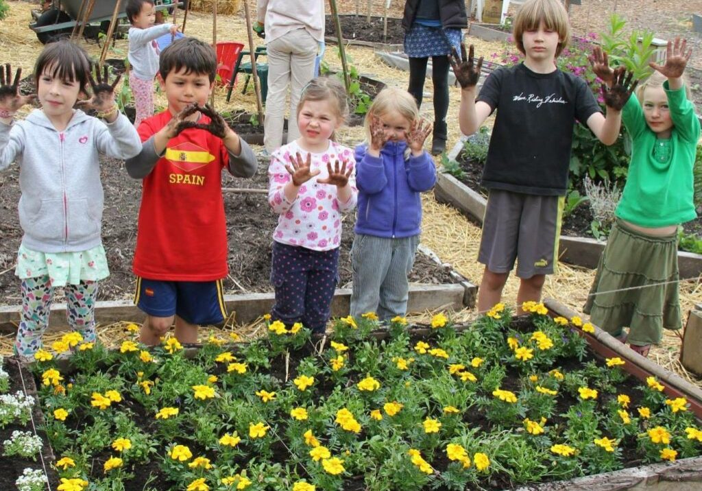 Children stand with muddy hands in a garden, showcasing a raised bed filled with yellow flowers. It's a delightful family workshop, where blooms flourish and young gardeners learn the joys of nurturing plants together.